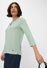Mintgroene top met 3/4 mouwen, voorzien van een sleutelgatenhals en zijsplitten. Zacht materiaal met subtiele details aan de onderkant.