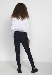 Marina stretchbyxor med en figurnära design och elastisk midja, kombinerat med en vit långärmad skjorta. Svarta skor med tjocka sulor.
