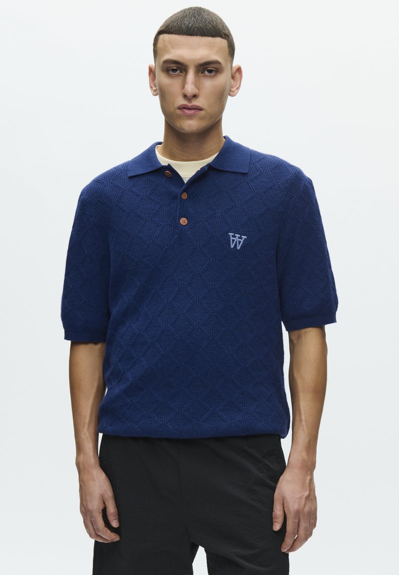 Polo bleu marine à texture en losanges, manches courtes, col et deux boutons marron. Présente un logo brodé discret sur la poitrine.