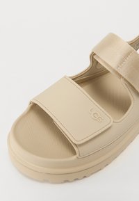 Beige gummisandal med teksturert såle og justerbar stropp. Har et logo preget på siden og en tykk, polstret såle.