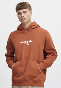 Sudadera color óxido hecha de tela suave, con un bolsillo frontal, que presenta un gráfico blanco de una figura y árboles en el pecho.