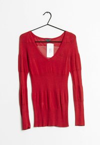 Červený žebrovaný top s dlouhými rukávy, výstřihem do V a mírně rozšířenými manžetami. Materiál je měkký s hladkou texturou, štítek viditelný na přední straně.