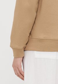 Sweatshirt bege com punhos e barra canelados, apresentando uma textura suave. Um tecido branco e em camadas aparece abaixo da linha da barra.