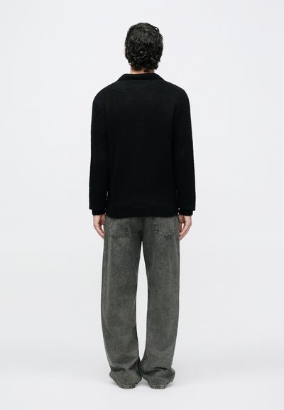 Bărbat cu păr creț, purtând un pulover negru texturat și blugi gri comozi, stând cu fața spre spate pe un fundal simplu alb.