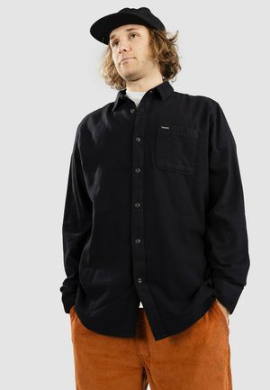 Camisa negra de botones, hecha de una tela texturizada, con un bolsillo en el pecho y un corte relajado, combinada con pantalones de pana naranja.