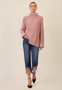 Rosa gestrickter Pullover mit hohem Kragen, kombiniert mit blauen, cropped Jeans mit Fransen am Saum und schwarzen spitzen High Heels.
