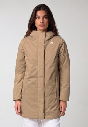 Donna che indossa un cappotto invernale beige con cappuccio e tasche frontali, in piedi davanti a uno sfondo chiaro e uniforme.
