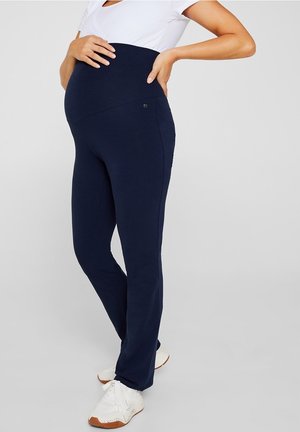 Pregnant person wearing navy blue maternity pants and a white short-sleeve top, standing with one hand on belly and one on lower back.