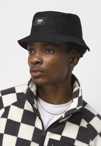 Black bucket hat with Vans logo patch. Model wears a black and white checkered fleece jacket, with a collar and zipper detail.