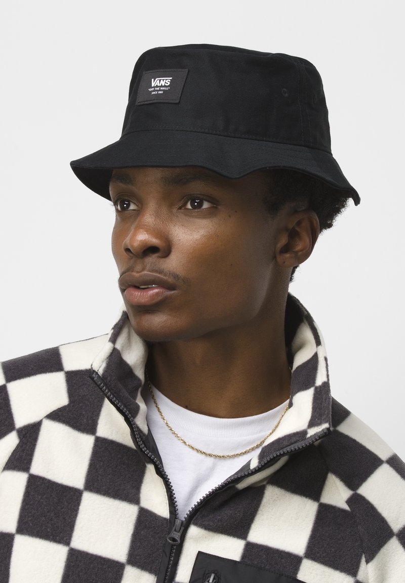 Black bucket hat with Vans logo patch. Model wears a black and white checkered fleece jacket, with a collar and zipper detail.
