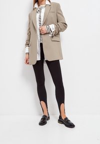 Blazer oversized beige, chemise blanche à rayures noires, leggings noirs avec fentes latérales et mocassins noirs avec accents jaunes.