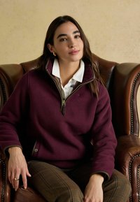 Jauna sieviete ar gariem tumšiem matiem sēž uz brūna ādas krēsla, ģērbusies baltā krekliņā ar apkakli, bordo flīsa džemperī un raibās biksēs.