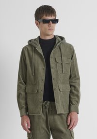Hombre vestido con chaqueta y pantalones de pana color verde olivo, camisa negra y gafas de sol rectangulares negras, de pie frente a un fondo gris simple.