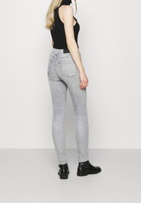 Mujer vestida con unos pantalones skinny grises claros, un top sin mangas negro y botas negras de tobillo, de pie contra un fondo blanco liso.