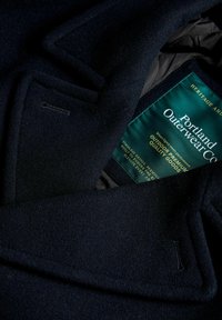 Marineblauwe wollen jas met een ingesneden kraag, klepzak en een groene merklabel aan de binnenkant. Zachte textuur met een klassiek ontwerp.