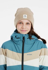 Smėlio spalvos megztas beanie su logotipo pachat, dėvimas su mėlyna ir smėlio spalvos chevron dizaino striuke, pagaminta iš lygaus audinio, su užtrauktuko uždarymu ir gobtuvu.
