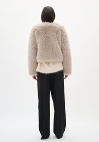 Chaqueta de piel sintética en beige claro, con un diseño corto y una textura esponjosa, superpuesta a una blusa suave en crema, combinada con pantalones negros holgados.