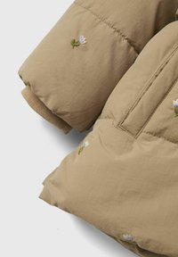 Doudoune tan rembourrée avec broderies florales ; elle possède un tissu doux, des poignets côtelés et une texture lisse. Coutures détaillées sur les épaules.