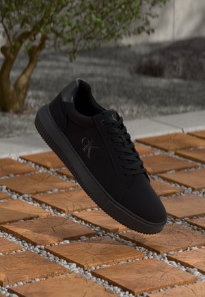 Black sneaker with smooth leather upper, round toe, and flat rubber sole. Features lace-up closure and embossed logo. Set on textured ground.
