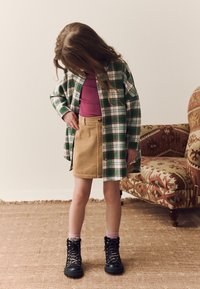 Grün-weiß kariertes Hemd mit Knöpfen, lila enganliegendes Top, beigefarbener Cordrock mit Knöpfen, schwarze Schnürstiefel, rosa Socken.