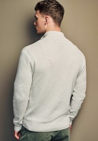 Pull à col en tricot texturé gris clair, avec des poignets et un ourlet côtelés. Le modèle est positionné de manière latérale contre un fond uni.