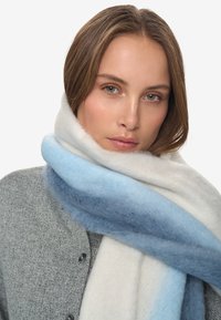 Jonge vrouw met lichtbruin haar draagt een dikke zachte sjaal in witte, lichtblauwe en blauwe tinten over een grijze gebuttonde trui.