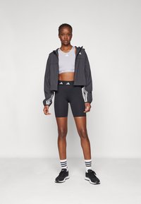 Kvinne som står i svarte treningsshorts, grå sports-BH, svart jakke med hvite striper, hvite sokker med svarte striper, og svarte joggesko.