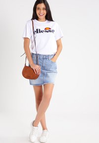Hvit kortermet t-skjorte med svart logo, kombinert med et lyseblått denimskjørt, hvite joggesko og en liten brun skinncrossbody-veske.