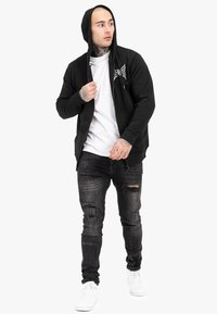 Sort lynlås hoodie med et logo, båret over en hvid t-shirt. Kombineret med slidte sorte jeans og hvide sneakers. Glat tekstur.