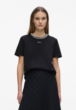 Kvinde iført en sort t-shirt med perlebesat krave og teksten "Karl", kombineret med et tekstureret sort skørt, stående mod en hvid baggrund.