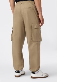 Beige cargobroek met elastische tailleband en enkels, met grote zijzakken, gedragen met witte Champion sneakers en een zwart top.