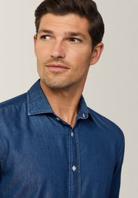 Camicia in denim blu scuro, con chiusura a bottoni, colletto a punta e dettagli di cucitura discreti. Tessuto morbido e vestibilità sagomata.