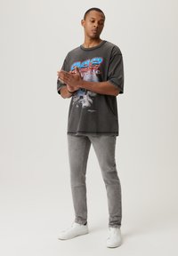 Szara koszulka graficzna z kolorowym wzorem z przodu, zestawiona z dopasowanymi szarymi dżinsami i białymi sneakersami. Luźny krój i materiał.