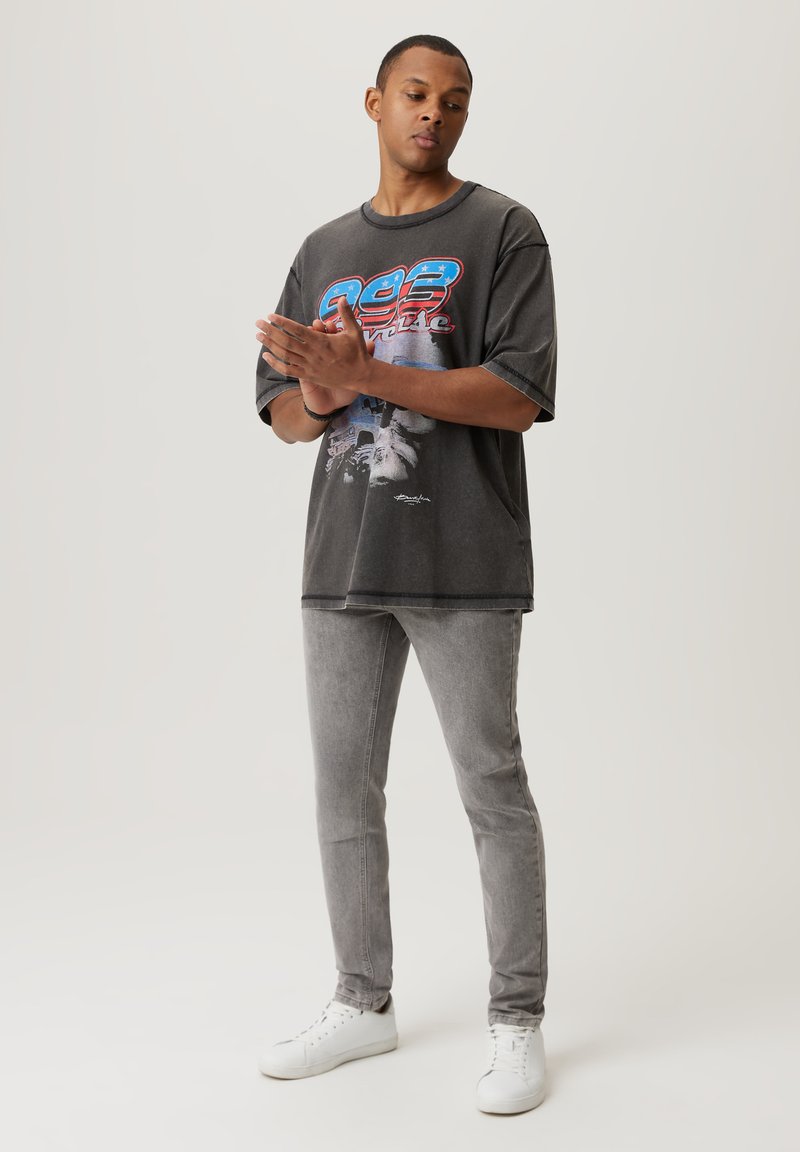 Szara koszulka graficzna z kolorowym wzorem z przodu, zestawiona z dopasowanymi szarymi dżinsami i białymi sneakersami. Luźny krój i materiał.