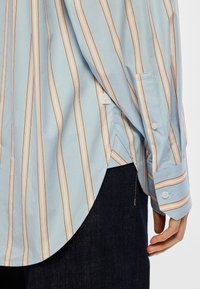 Ανοιχτό μπλε πουκάμισο με πορτοκαλί και λευκές κάθετες ρίγες. Διαθέτει κουμπωτές μανσέτες και πλαϊνή σχισμή για ευκολία κινήσεων. Φτιαγμένο από βαμβακερό υλικό.