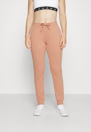 Jogging beige slim fit avec taille à cordon, porté avec un soutien-gorge de sport blanc et des baskets blanches, modèle debout devant un fond uni.