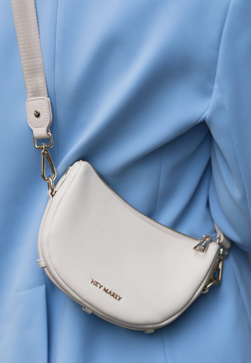 Sac en cuir blanc en bandoulière avec une forme courbée, des accents en matériel doré et le nom de la marque embossé, porté contre un vêtement bleu clair.
