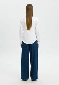 Camicia bianca a maniche lunghe con polsini abbottonati, abbinata a jeans in denim blu a gamba larga. I jeans hanno un taglio alto e una leggera usura.