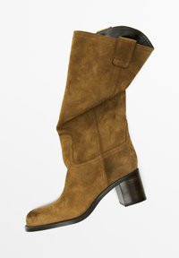 Stivale in suede marrone fino al ginocchio con parte superiore morbida, punta arrotondata e tacco in legno impilato. Dettagli sulle cuciture laterali e fodera interna nera.