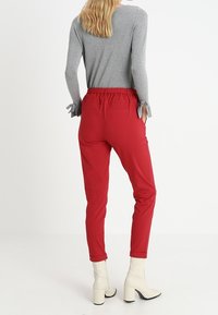 Røde tapered bukser med elastisk taljebånd, forlommer og en glat tekstur, parret med en grå langærmet bluse med sløjfe detaljer.
