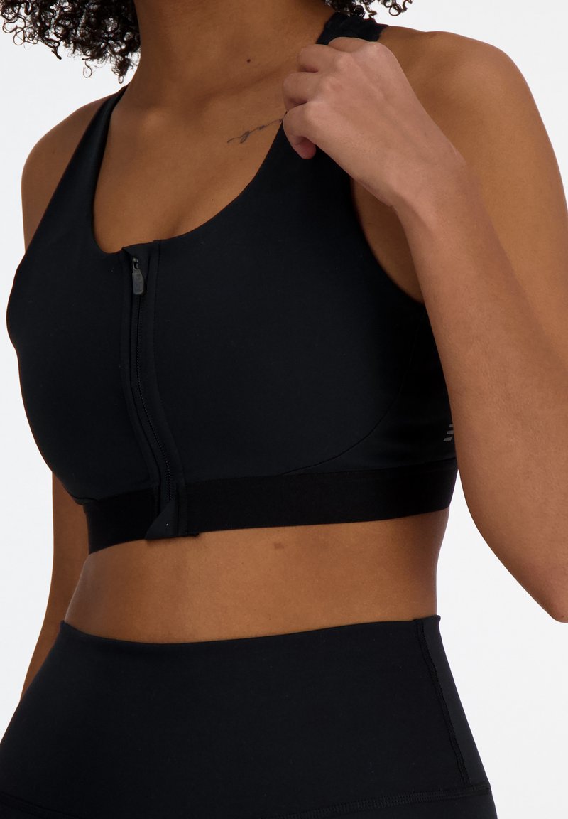 Soutien-gorge de sport noir en tissu lisse et extensible, avec une fermeture éclair à l'avant et une large bande élastique en bas pour le maintien.