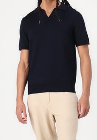 Marinblå kortärmad polo med V-ringning, med textilstruktur och ribbad nederkant, tillsammans med ljusa byxor.