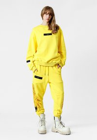 Sudadera de algodón amarilla con etiqueta negra y joggers a juego que presentan un estampado floral en la pierna izquierda. Combinado con botas blancas con cordones.