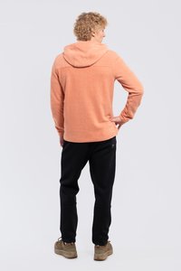 Mand står med hånden på hoften, iført en ferskenfarvet hættetrøje i fleece, sorte bukser og beige vandresko på en ensfarvet baggrund.