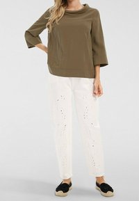 Haut vert olive à col bénitier et manches trois-quarts, associé à un pantalon blanc avec des détails de découpe en forme de feuilles. Chaussures espadrilles noires.