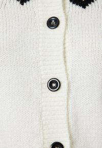 Cárdigan blanco de punto con un diseño acanalado y botones negros. El diseño incluye una textura vertical y un acento oscuro contrastante en la parte superior.