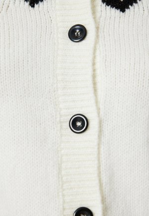 Cardigan bianco lavorato a maglia con design a coste e bottoni neri. Il design include una texture verticale e un accento scuro a contrasto nella parte superiore.