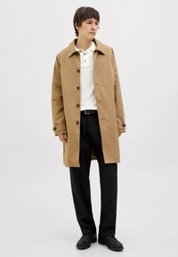 Hnědý bavlněný trench coat s širokým límcem, zdobený knoflíkovým zapínáním a bočními kapsami. Nositý přes bílou polokošili a černé kalhoty.