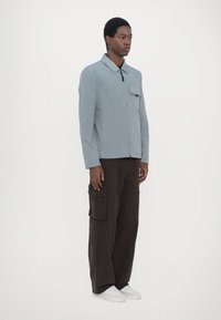 Lys grå skjortejakke med krave og en brystlomme, båret med mørkebrune cargos og hvide sneakers. Simpelt design, afslappet pasform.