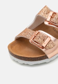 Rosaguld flat sandal med två glittrande remmar och metalliska spännen. Korkfotbädd och vit, strukturerad yttersula. Slät innervaddering.